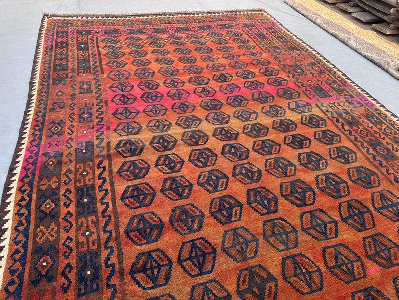 May include: A large, rectangular rug with a geometric pattern. The rug features a gradient of colours, transitioning from a vibrant pink to orange and brown. Dark blue geometric shapes are evenly spaced across the rug's surface. The border has intricate patterns in various colours.