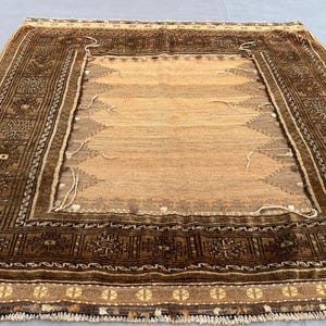 Puede incluir: Una alfombra rectangular tejida a mano con un campo central de color beige y un borde marrón oscuro. El borde presenta intrincados patrones geométricos y motivos florales. La alfombra tiene un borde con flecos y un aspecto vintage.