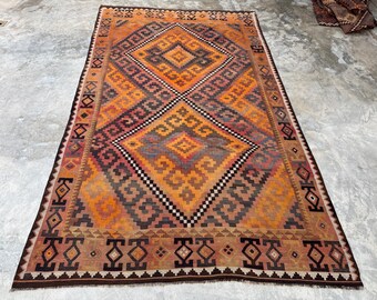 May include: A rectangular, hand-woven rug with a geometric pattern in shades of orange, brown, and gray. The central design features a diamond shape with smaller diamond and zigzag patterns. The rug has a dark brown border.