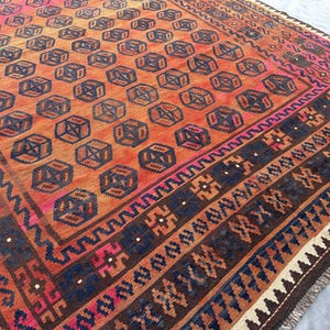 May include: A large, rectangular rug with a geometric pattern. The rug features a repeating pattern of dark blue cube-like shapes on an orange background. Borders include dark blue, brown, and pink accents. The rug has a fringed edge.