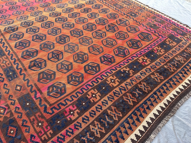 May include: A large, rectangular rug with a geometric pattern. The rug features a repeating pattern of dark blue cube-like shapes on an orange background. Borders include dark blue, brown, and pink accents. The rug has a fringed edge.