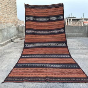 May include: A long, rectangular rug with a striped pattern in shades of brown, black, and white. The rug has a traditional, woven look and is made of natural fibers.