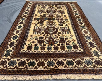 Puede incluir: Alfombra rectangular beige con un patrón geométrico central en marrón y marrón oscuro. La alfombra tiene un borde con diseños florales y geométricos repetidos en marrón, azul y verde. Tiene flecos.