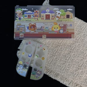 Könnte beinhalten: Eine durchscheinende Schutzhülle für eine Spielkonsole und zwei Controller-Cover mit Animal-Crossing-Figuren. Die Konsolenrückseite zeigt eine Stadtszene mit K.K. Slider, während die Controller-Cover verschiedene Spielsymbole und Charaktere wie Raymond und Gulliver abbilden.