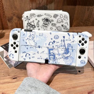 May include: A white gaming console case featuring blue cat illustrations, hearts, and text including "lovely kitty" and "party time." Behind it sits a white carrying case decorated with black line art of an astronaut and space elements.