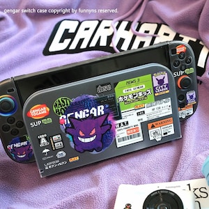 May include: A grey gaming console protective case featuring a variety of Gengar-themed stickers, including Japanese text, warning labels, and a prominent purple Gengar character graphic. The case is decorated with multiple colorful decals.