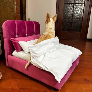 Può includere: Un piccolo letto rosa per cani con testiera trapuntata e dettagli in cristallo. Il letto include un cuscino bianco e una coperta bianca. Un piccolo cane con pelo marrone e bianco è seduto sul letto.