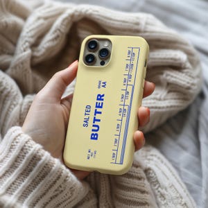 May include: A light yellow phone case with a blue design that mimics a stick of salted butter. The case features the words "SALTED BUTTER" and measurement markings for tablespoons and cups. The net weight is 4 oz (113g).