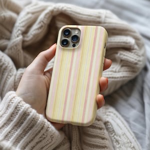 May include: A yellow phone case with vertical stripes in pink and white. The case has a camera module with three lenses. The phone case is held in a hand, with a neutral-colored sweater in the background.