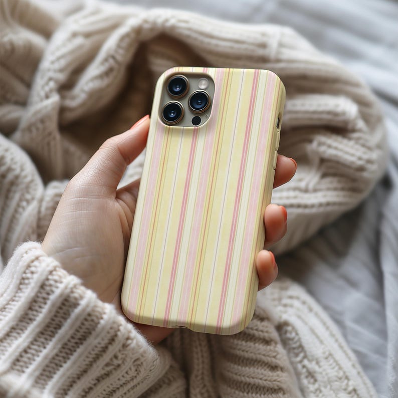 May include: A yellow phone case with vertical stripes in pink and white. The case has a camera module with three lenses. The phone case is held in a hand, with a neutral-colored sweater in the background.