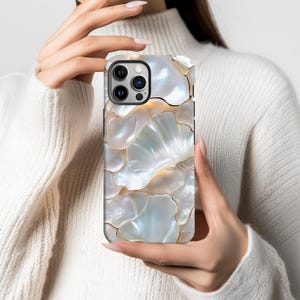 May include: A phone case with a mother-of-pearl effect and a floral design. The case has a pattern of overlapping, iridescent petal-like shapes in white, cream, and gold. The phone case has a black border and a camera cutout.