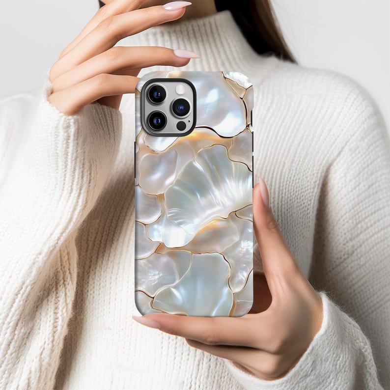 May include: A phone case with a mother-of-pearl effect and a floral design. The case has a pattern of overlapping, iridescent petal-like shapes in white, cream, and gold. The phone case has a black border and a camera cutout.