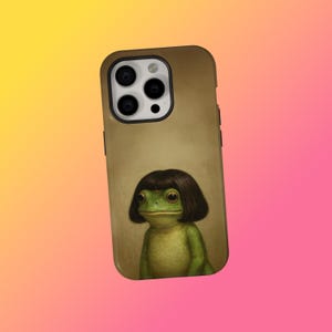 Puede incluir: Funda de teléfono con una rana verde con un corte de pelo bob marrón oscuro. La rana tiene ojos grandes y está sobre un fondo beige. La funda tiene un borde marrón oscuro y un recorte para la cámara.