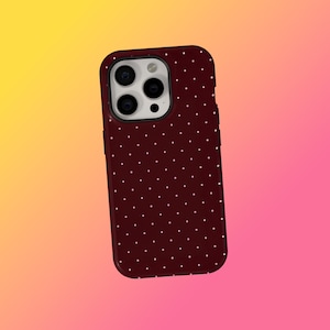 May include: A maroon phone case featuring a white polka dot design. The case has a shiny surface and a camera aperture. The background is a gradient of yellow, orange, and pink.