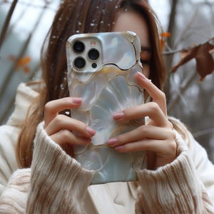 May include: A light blue and white phone case with a shell-like pattern. The case has gold-coloured accents. The phone case is held in front of a blurred background.