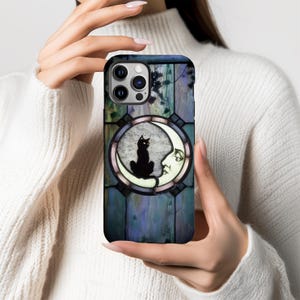 Puede incluir: Una funda de tel&eacute;fono con un dise&ntilde;o de vidriera. Presenta un gato negro sobre una luna creciente, sobre un fondo azul oscuro y morado con un efecto de panel de madera. El gato y la luna est&aacute;n en un marco circular.