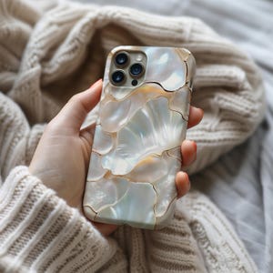 May include: A phone case with a mother-of-pearl effect, featuring a mosaic design. The case has a pattern of iridescent, shell-like shapes in white, cream, and gold. The phone case is held in a hand, with a soft, neutral-coloured sweater in the background.