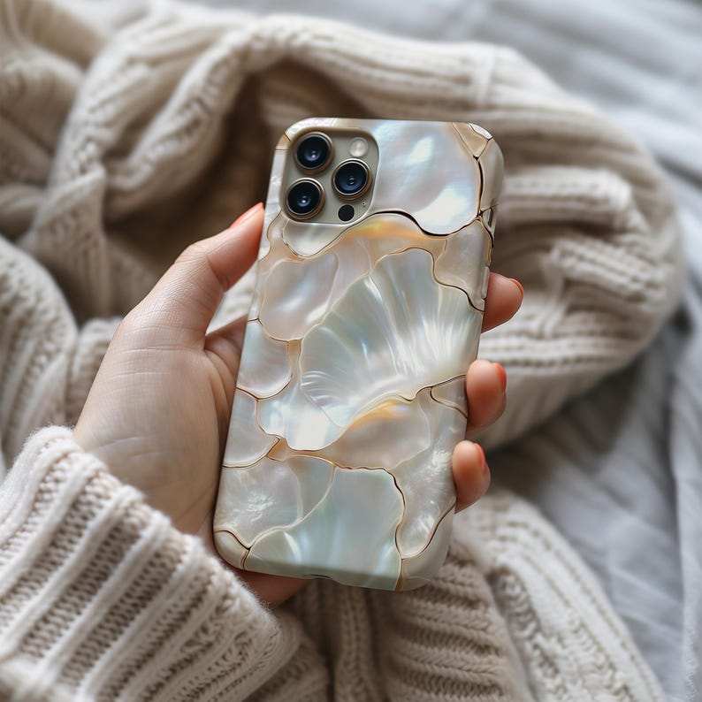 May include: A phone case with a mother-of-pearl effect, featuring a mosaic design. The case has a pattern of iridescent, shell-like shapes in white, cream, and gold. The phone case is held in a hand, with a soft, neutral-coloured sweater in the background.