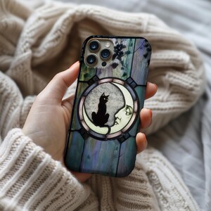 Puede incluir: Una funda de tel&eacute;fono con un dise&ntilde;o de vidriera que representa un gato negro sobre una luna creciente. La funda tiene un fondo azul, verde y morado con un borde decorativo. La funda del tel&eacute;fono la sostiene una persona.