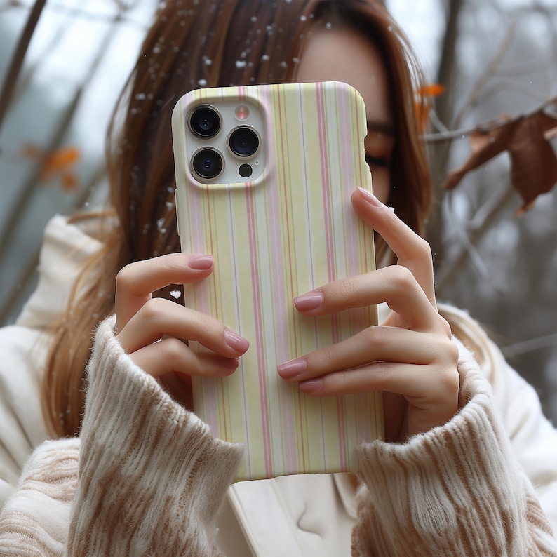 May include: A yellow and pink striped phone case with a camera lens cutout. The case is held in front of a person, with a blurred background of a forest. The case has a vertical stripe pattern.