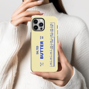 May include: A light yellow phone case featuring a design that mimics a stick of salted butter. The case has blue text that reads "SALTED BUTTER AA" and includes a measurement conversion chart. The net weight is listed as 4 oz (113g).