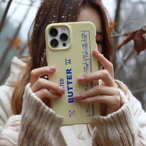 May include: A light yellow phone case with the word "BUTTER" in blue and white text. The case has a butter-themed design with measurements in tablespoons and cups. The case also has the text "NET WT. 4 OZ (113G)" and "GRADE AA".