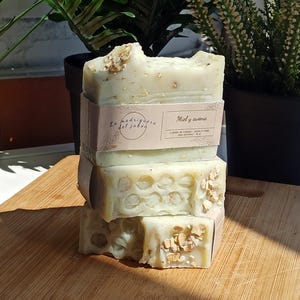 Può includere: Tre saponette fatte a mano impilate su una superficie di legno. Il sapone è verde e bianco con un motivo a nido d'ape e fiocchi di avena. L'etichetta recita "La Madriguera del Jabón" e "Miel y avena".