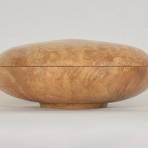 May include: A wooden bowl with a natural wood finish. The bowl is round and has a slightly raised base. The wood grain is visible and has a light brown color.