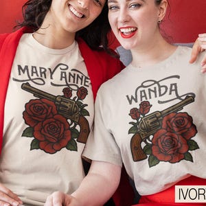 Pode incluir: T-shirts cor marfim com um design gráfico com um revólver, rosas vermelhas e os nomes "Mary Anne" e "Wanda" em escrita preta. As t-shirts são usadas por duas pessoas. A palavra "IVORY" é exibida na parte inferior.