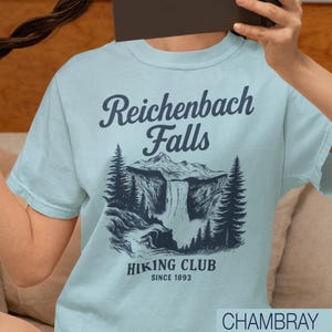 May include: Light blue t-shirt with a graphic of Reichenbach Falls, a mountain, and trees. The text "Reichenbach Falls" is above the graphic, and "Hiking Club Since 1893" is below. The shirt is chambray.