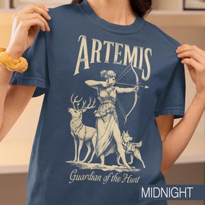 May include: Midnight blue t-shirt featuring a graphic of Artemis, the Greek goddess of the hunt, with a bow and arrow, a deer, and a dog. The text "ARTEMIS" and "Guardian of the Hunt" are displayed in a vintage style.