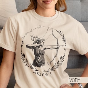 May include: Ivory t-shirt with a black and white illustration of Artemis, the Greek goddess of the hunt, aiming a bow and arrow. The design includes the text "The Huntress Rises" and the name "ARTEMIS".