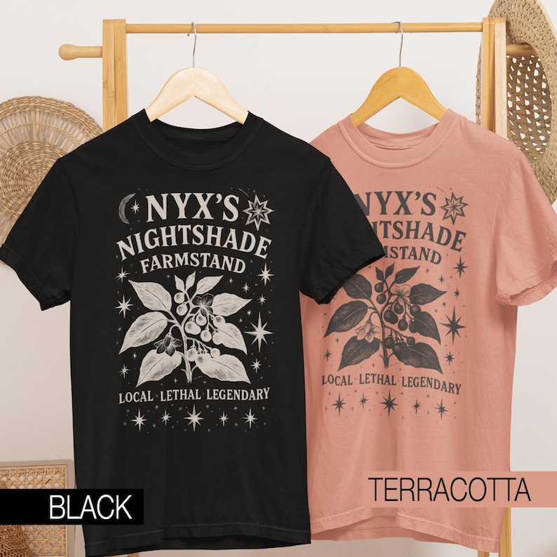 May include: Two t-shirts, one black and one terracotta, hanging on a wooden rack. Each shirt features a graphic with the text "NYX'S NIGHTSHADE FARMSTAND" and a botanical illustration. The phrase "LOCAL LETHAL LEGENDARY" is also printed on the shirts.