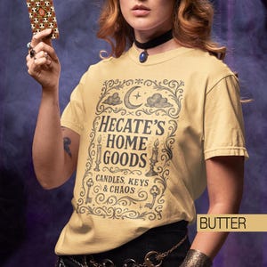 May include: Pale yellow t-shirt featuring the words "Hecate's Home Goods" in an ornate design with candles, keys, and a crescent moon. A person holds a patterned card. The shirt is a light, neutral colour.