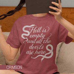Puede incluir: Camiseta color carmesí con la frase "Hell is empty and all the devils are here" en escritura blanca, acompañada de ilustraciones de serpientes. La palabra "CRIMSON" se muestra en la parte inferior.