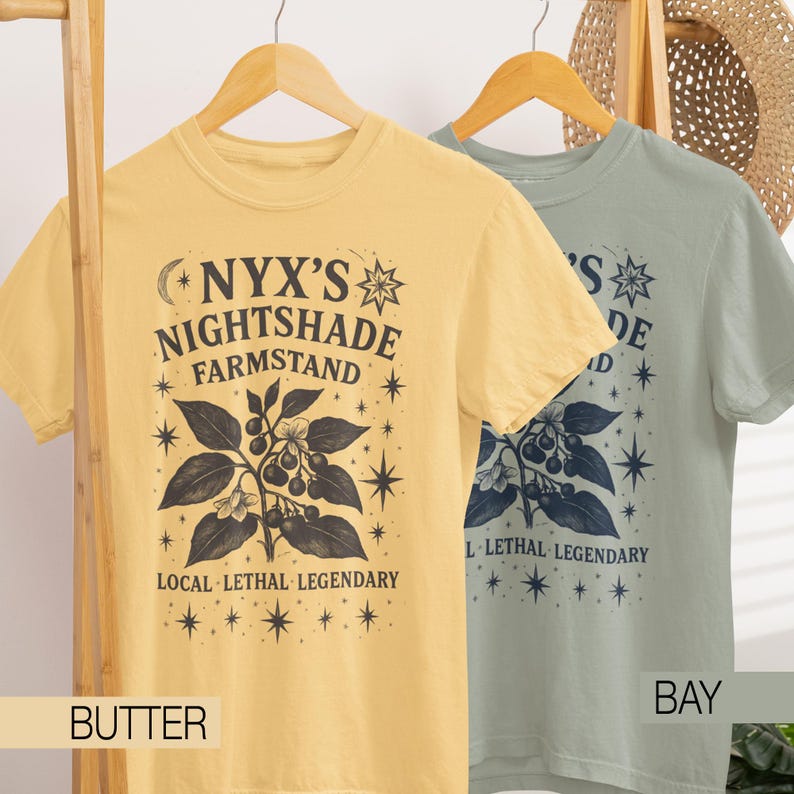 May include: Two short-sleeved t-shirts, one butter yellow and one bay green, hang on wooden hangers. The shirts feature a botanical illustration with the text "NYX'S NIGHTSHADE FARMSTAND" and "LOCAL LETHAL LEGENDARY". The shirts are a comfortable fit.