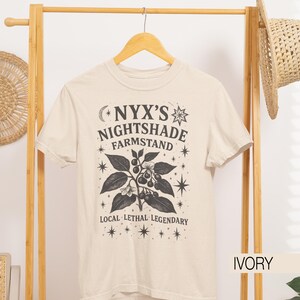 May include: Ivory-colored t-shirt with a graphic design featuring the text "NYX'S NIGHTSHADE FARMSTAND" and a botanical illustration of a plant with berries and leaves. The text "LOCAL LETHAL LEGENDARY" is also included.