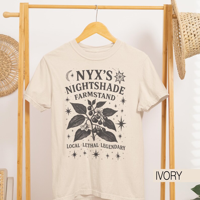 May include: Ivory-colored t-shirt with a graphic design featuring the text "NYX'S NIGHTSHADE FARMSTAND" and a botanical illustration of a plant with berries and leaves. The text "LOCAL LETHAL LEGENDARY" is also included.