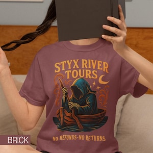 May include: Brick-colored t-shirt with the text "STYX RIVER TOURS" and an illustration of a hooded figure in a boat. The design includes a crescent moon and stars, with the words "NO REFUNDS - NO RETURNS" below the graphic.