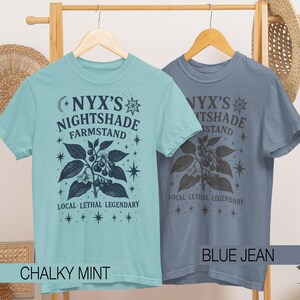 May include: Two short-sleeved t-shirts, one chalky mint and one blue jean, hang on a wooden rack. Each shirt features the text "NYX'S NIGHTSHADE FARMSTAND" with a botanical illustration and the words "LOCAL LETHAL LEGENDARY".
