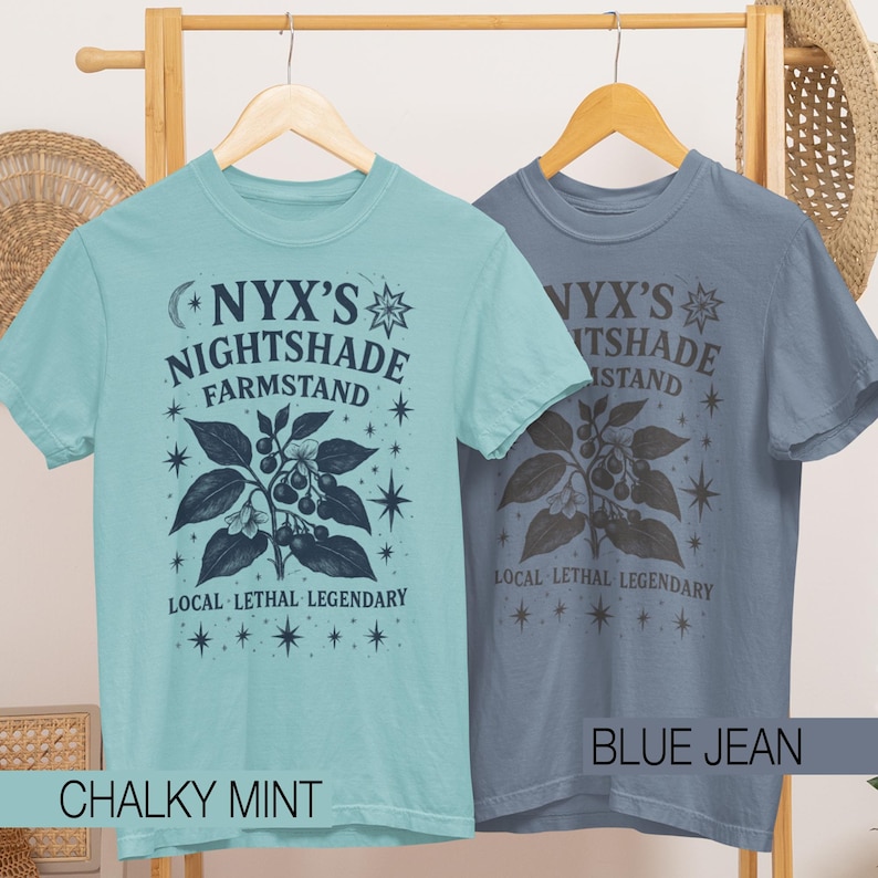 May include: Two short-sleeved t-shirts, one chalky mint and one blue jean, hang on a wooden rack. Each shirt features the text "NYX'S NIGHTSHADE FARMSTAND" with a botanical illustration and the words "LOCAL LETHAL LEGENDARY".
