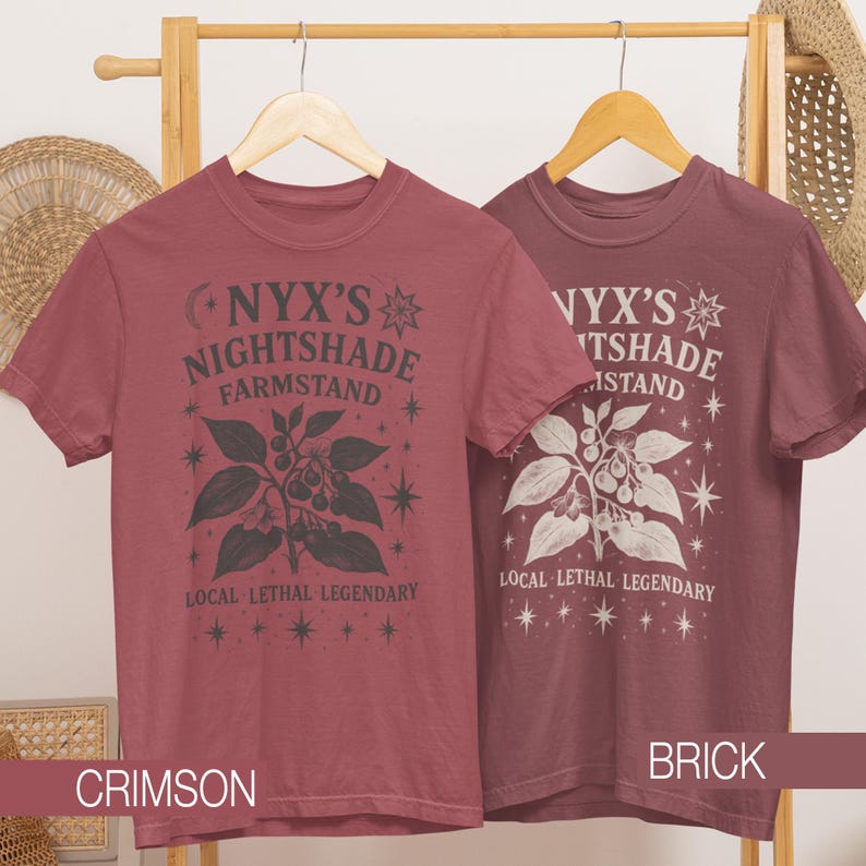 May include: Two short-sleeved t-shirts in crimson and brick colors, hanging on a wooden rack. Each shirt features a graphic with the text "NYX'S NIGHTSHADE FARMSTAND" and a botanical illustration. The text "LOCAL LETHAL LEGENDARY" is also printed on the shirts.