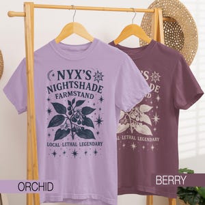 May include: Two short-sleeved t-shirts, one orchid and one berry, hang on a wooden rack. Each shirt features a graphic with the text "NYX'S NIGHTSHADE FARMSTAND" and botanical illustrations. The phrase "LOCAL LETHAL LEGENDARY" is also printed on the shirts.