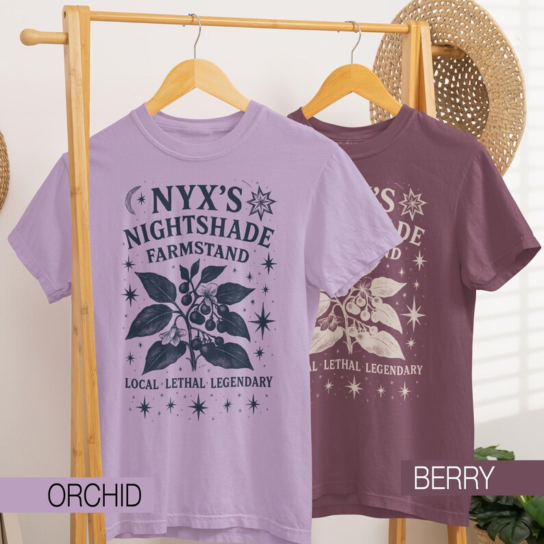 May include: Two short-sleeved t-shirts, one orchid and one berry, hang on a wooden rack. Each shirt features a graphic with the text "NYX'S NIGHTSHADE FARMSTAND" and botanical illustrations. The phrase "LOCAL LETHAL LEGENDARY" is also printed on the shirts.