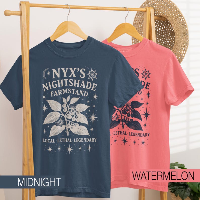 May include: Two short-sleeved t-shirts hang on a wooden rack. One is midnight blue, the other watermelon pink. Both shirts feature a white graphic with the text "NYX'S NIGHTSHADE FARMSTAND" and "LOCAL LETHAL LEGENDARY". The design includes botanical illustrations and stars.