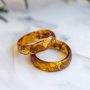 Könnte beinhalten: Zwei goldene Harzringe mit eingebetteten Blattgold. Die Ringe liegen übereinander auf einer weißen Marmorfläche.