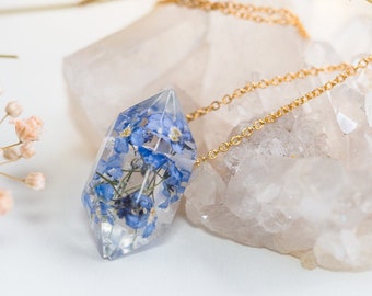 Può includere: Una collana con catena dorata con un ciondolo di cristallo trasparente che contiene fiori di nontiscordardime blu pressati.