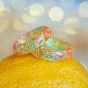 May include: Two colourful resin rings with glitter and iridescent flakes rest on a bright yellow lemon. The rings feature shades of orange, green, and gold, creating a shimmering effect. The background is a soft, out-of-focus bokeh.