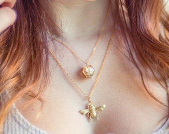 Può includere: Due collane d'oro. La collana superiore ha un ciondolo esagonale con un design floreale. La collana inferiore presenta un ciondolo a forma di ape dettagliato. La modella indossa una canotta grigia. Il marchio "13th Psyche" è visibile in basso.
