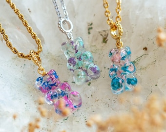 May include: Three gold and silver necklaces with gummy bear pendants. Each pendant is filled with pressed flowers in shades of blue, pink, and green.